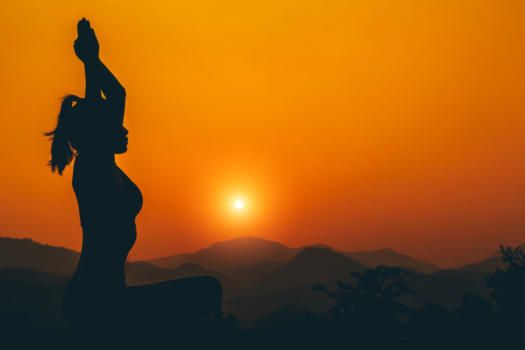 Na imagem aparece a silhueta de uma mulher praticando ioga com um fundo de pôr do sol laranja